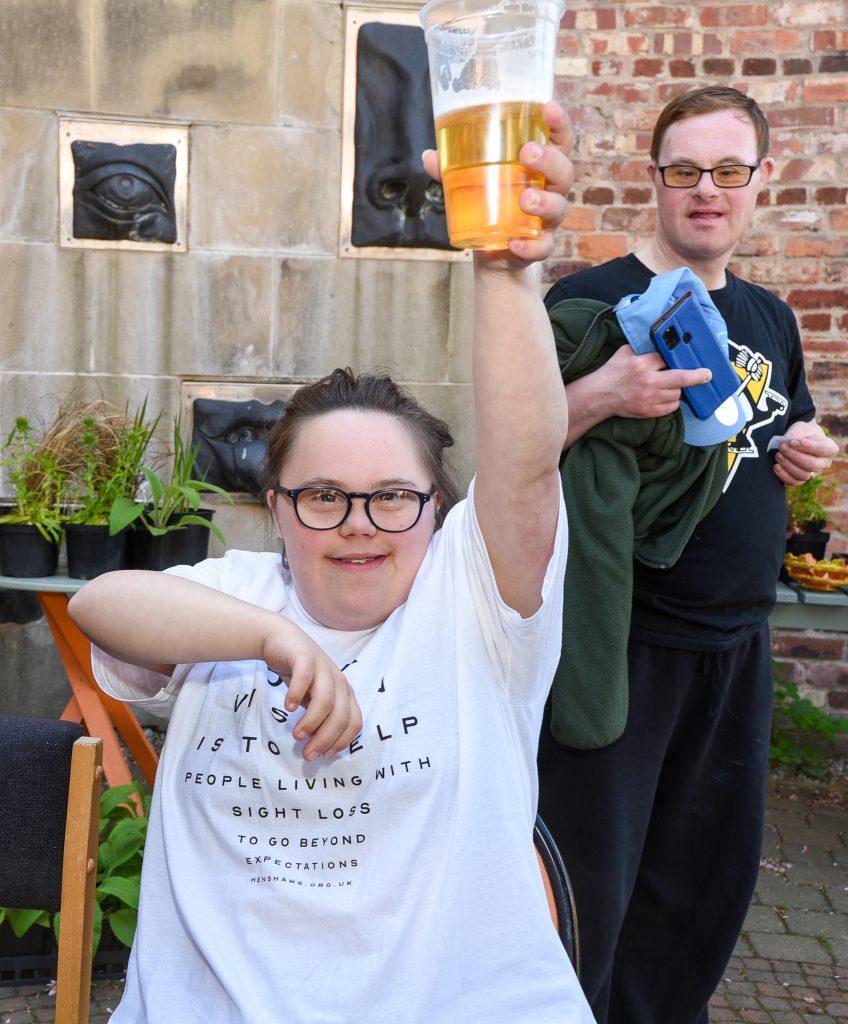 Photo of Henshaws art-maker Ebonie, raising a glass at the Henshaws Beer Festival in 2022, with another Henshaws service-user in the background.
