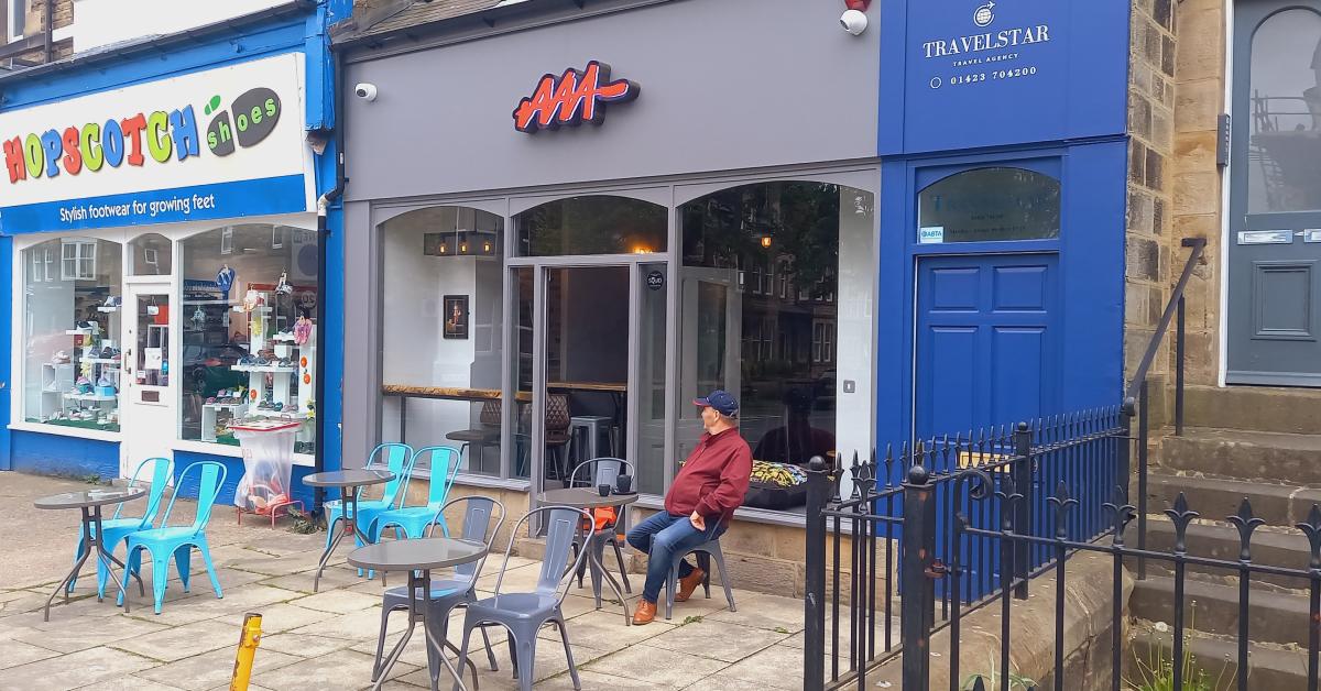 Photo of the exterior of AAA on Cold Bath Road, showing a customer seated at one of the four outdoor tables, with neighbouring businesses Hopscotch and Travelstar also in view.