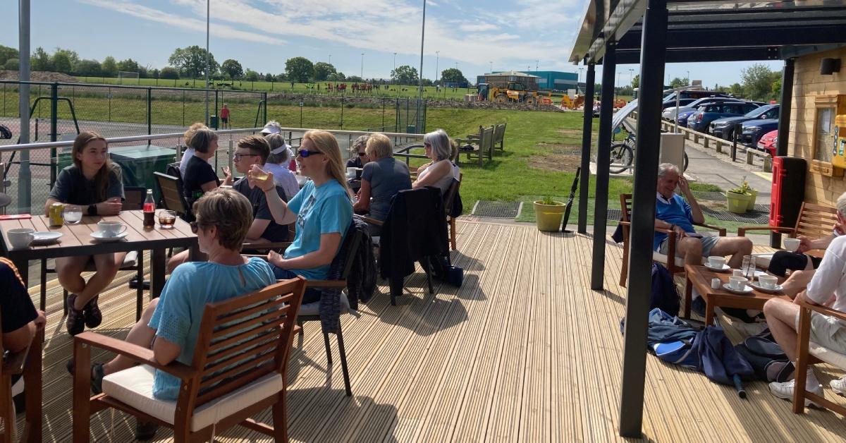 Photo of people sitting in the sun and drinking at café-style tables on the new terrace at Harrogate Spa Tennis Club.