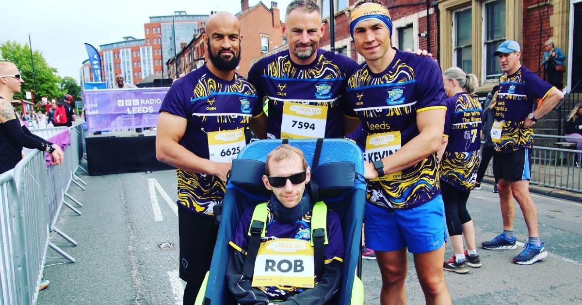 Photo of MND campaigner Rob Burrow in a wheelchair, with Jamie Jones-Buchanan (left) and Kevin Sinfield OBE behind him.