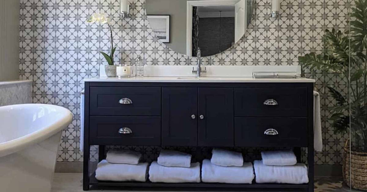 A lifestyle shot of a large navy bathroom vanity, with a mirror and a pattern tile on the walls