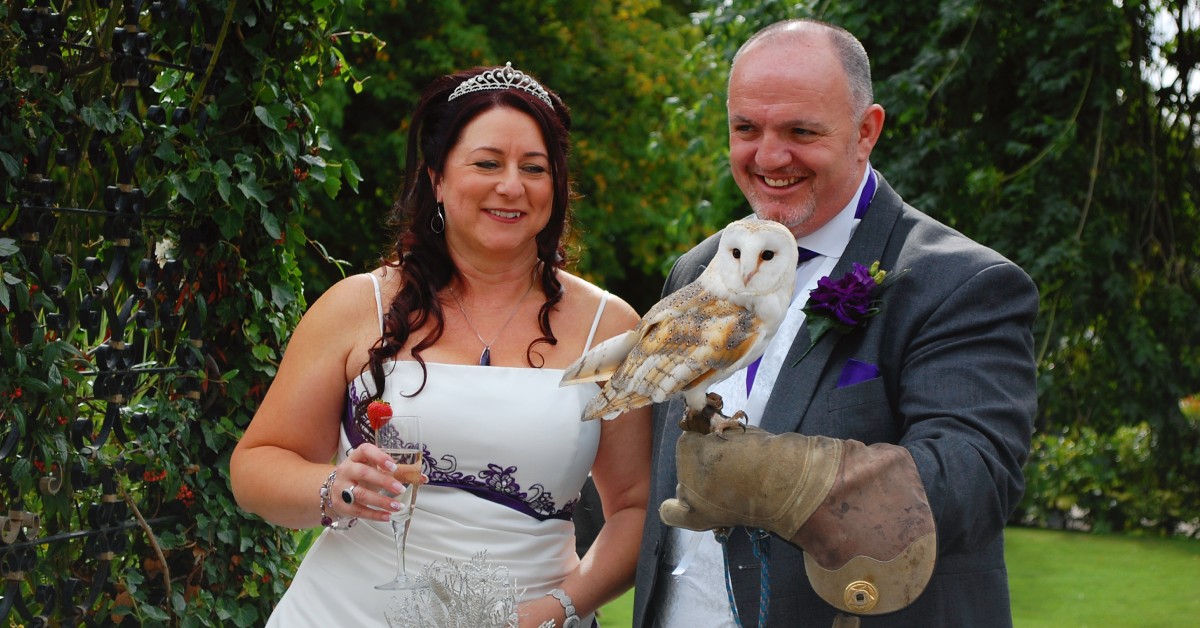 Photo of a happy bridge and groom, who is wearing a falconry gautlet and a white barn owl is perching on his hand.